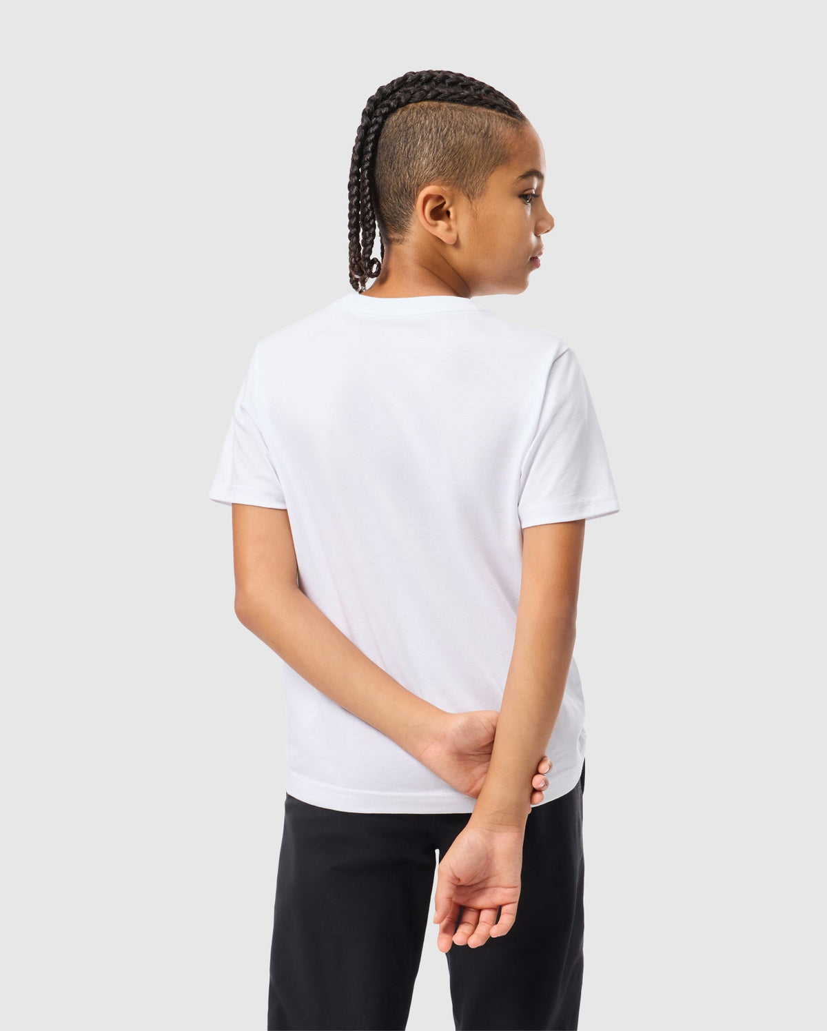 A young person with braided hair stands with their back to the camera, wearing a plain Kids Kingston Graphic Tee - B0U743D200 by Psycho Bunny in soft Pima cotton and black pants. Their right hand is held behind their back, set against a light gray background.