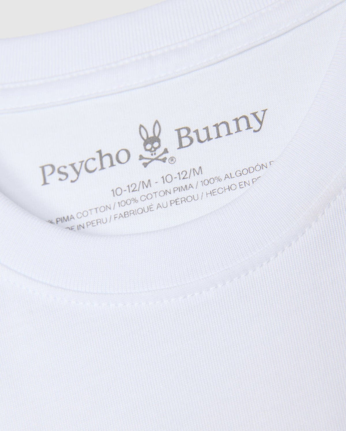 An up-close look at the neck label of the KIDS KINGSTON GRAPHIC TEE - B0U743D200, featuring the "Psycho Bunny" brand name next to a bunny with crossbones graphic. The label displays the size "10-12/M" and fabric information, emphasizing its premium 100% Pima cotton material, crafted in Peru.