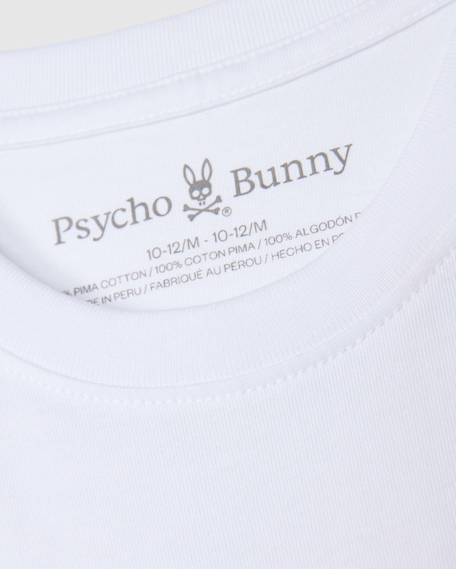 An up-close look at the neck label of the KIDS KINGSTON GRAPHIC TEE - B0U743D200, featuring the "Psycho Bunny" brand name next to a bunny with crossbones graphic. The label displays the size "10-12/M" and fabric information, emphasizing its premium 100% Pima cotton material, crafted in Peru.