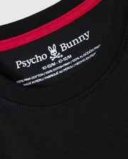 Close-up of a black KIDS MALTON GRAPHIC TEE - B0U795D200 collar with red trim. The label inside the collar displays the Psycho Bunny brand name and a bunny logo, along with size details "10-12/M" and material composition "100% Pima cotton," showcasing Peruvian craftsmanship in both English and Spanish.