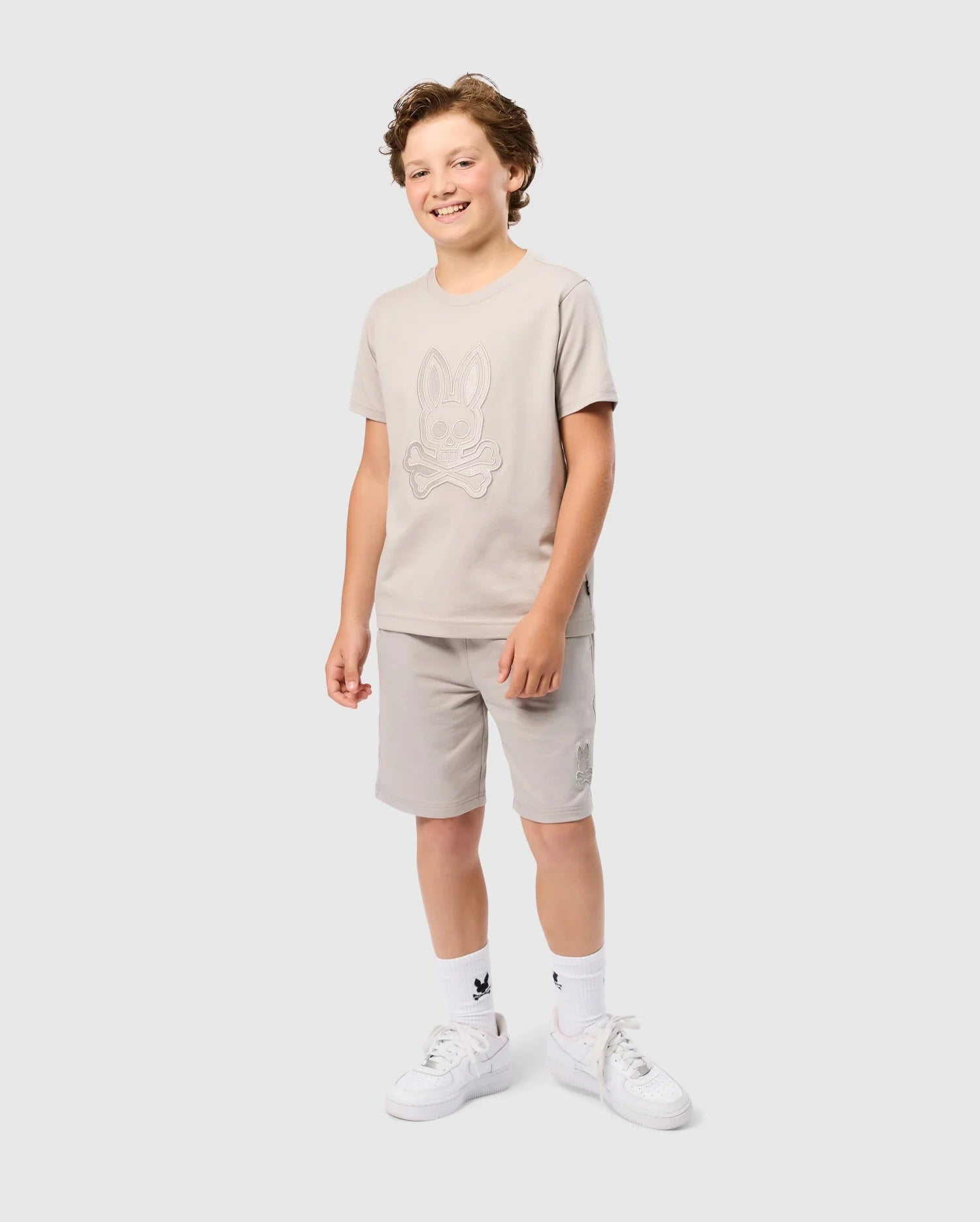 A young boy with curly hair is smiling and standing against a plain background. He is wearing a light beige Psycho Bunny Kids Malton Graphic Tee (B0U795D200) made from Pima cotton, matching shorts, white socks with a small graphic, and regular fit white sneakers. His hands are by his sides.