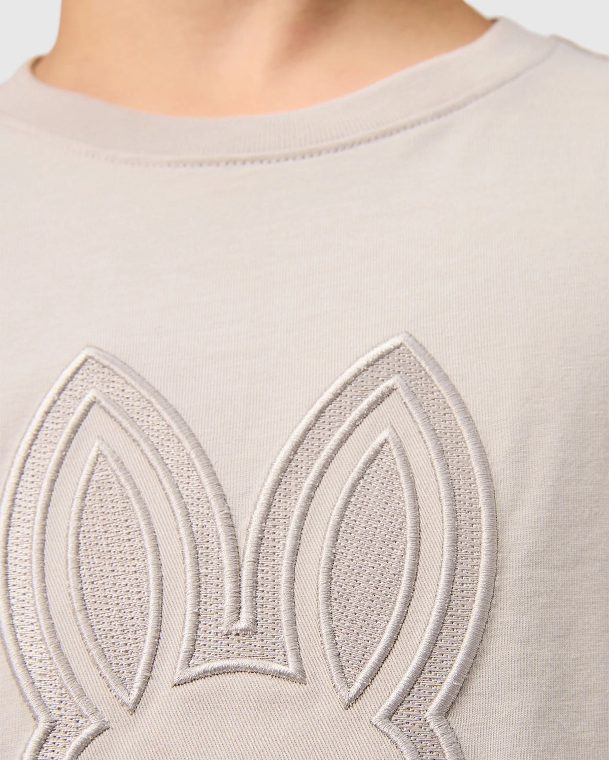 Close-up of the upper part of a Psycho Bunny KIDS MALTON GRAPHIC TEE - B0U795D200, featuring an embossed outline of rabbit ears on the chest area. The gray Pima cotton fabric appears smooth and soft, with detailed stitching highlighting the design. The background is plain, and part of a person's neck is visible at the top.