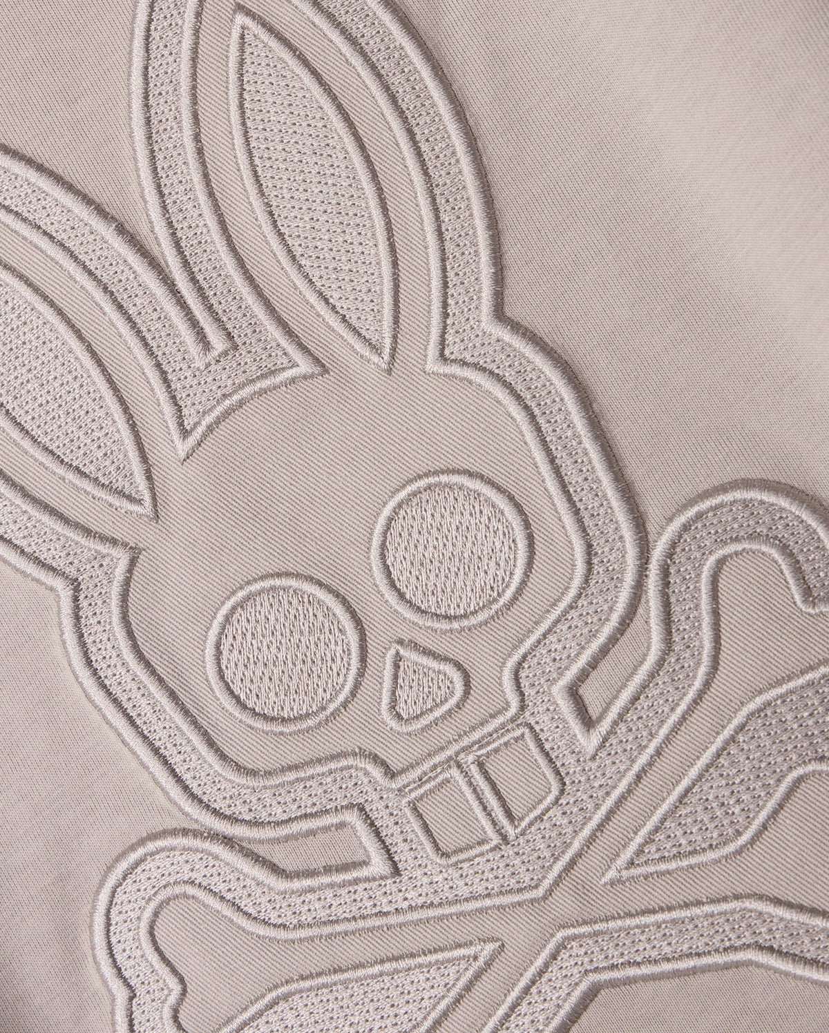 Close-up of a light-colored Pima cotton fabric with an intricate stitched design featuring a stylized bunny head with large ears and crossbones underneath. The finely detailed stitching on this Psycho Bunny Kids Malton Graphic Tee (B0U795D200) adds texture and depth to the design.