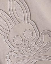 Close-up of a light-colored Pima cotton fabric with an intricate stitched design featuring a stylized bunny head with large ears and crossbones underneath. The finely detailed stitching on this Psycho Bunny Kids Malton Graphic Tee (B0U795D200) adds texture and depth to the design.