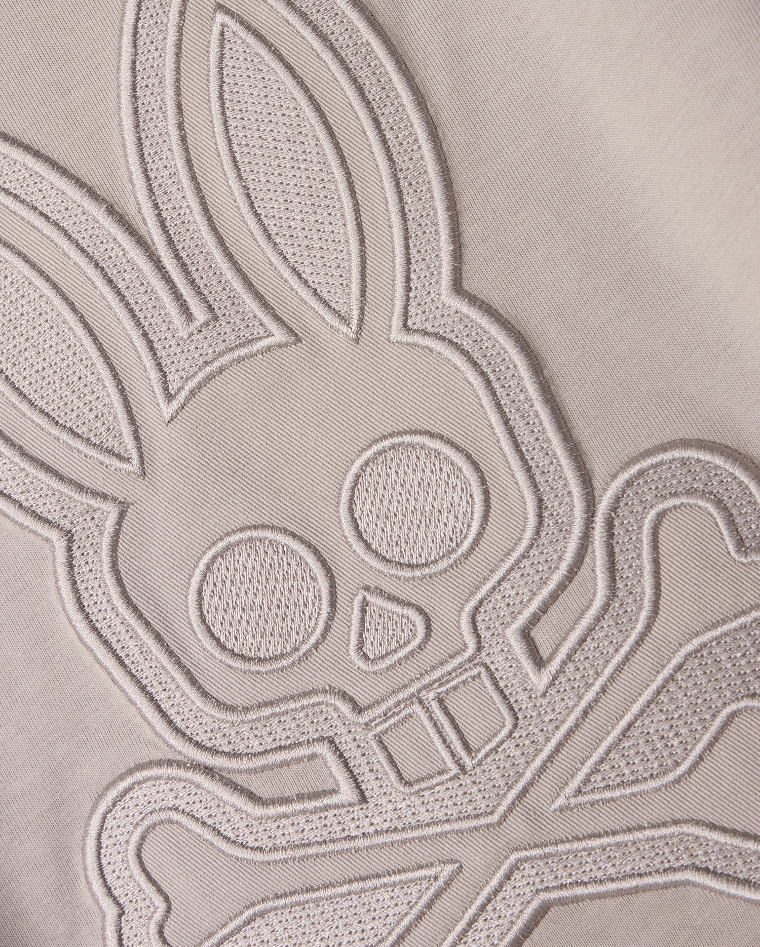 Close-up of a light-colored Pima cotton fabric with an intricate stitched design featuring a stylized bunny head with large ears and crossbones underneath. The finely detailed stitching on this Psycho Bunny Kids Malton Graphic Tee (B0U795D200) adds texture and depth to the design.