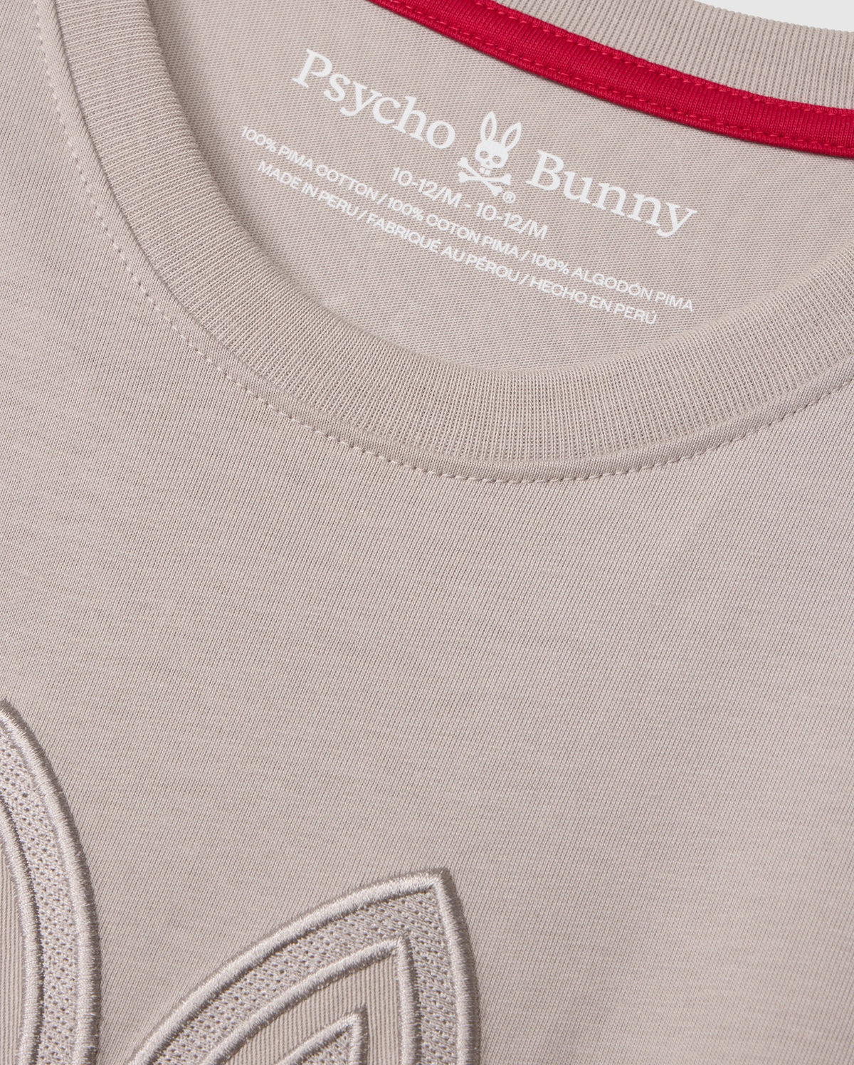 Close-up of a light-colored KIDS MALTON GRAPHIC TEE - B0U795D200 by Psycho Bunny, crafted from soft Pima cotton, featuring the brand's label and size details printed inside the collar. The collar is accented with a red lining, and part of an embroidered design is visible near the bottom left of the image.