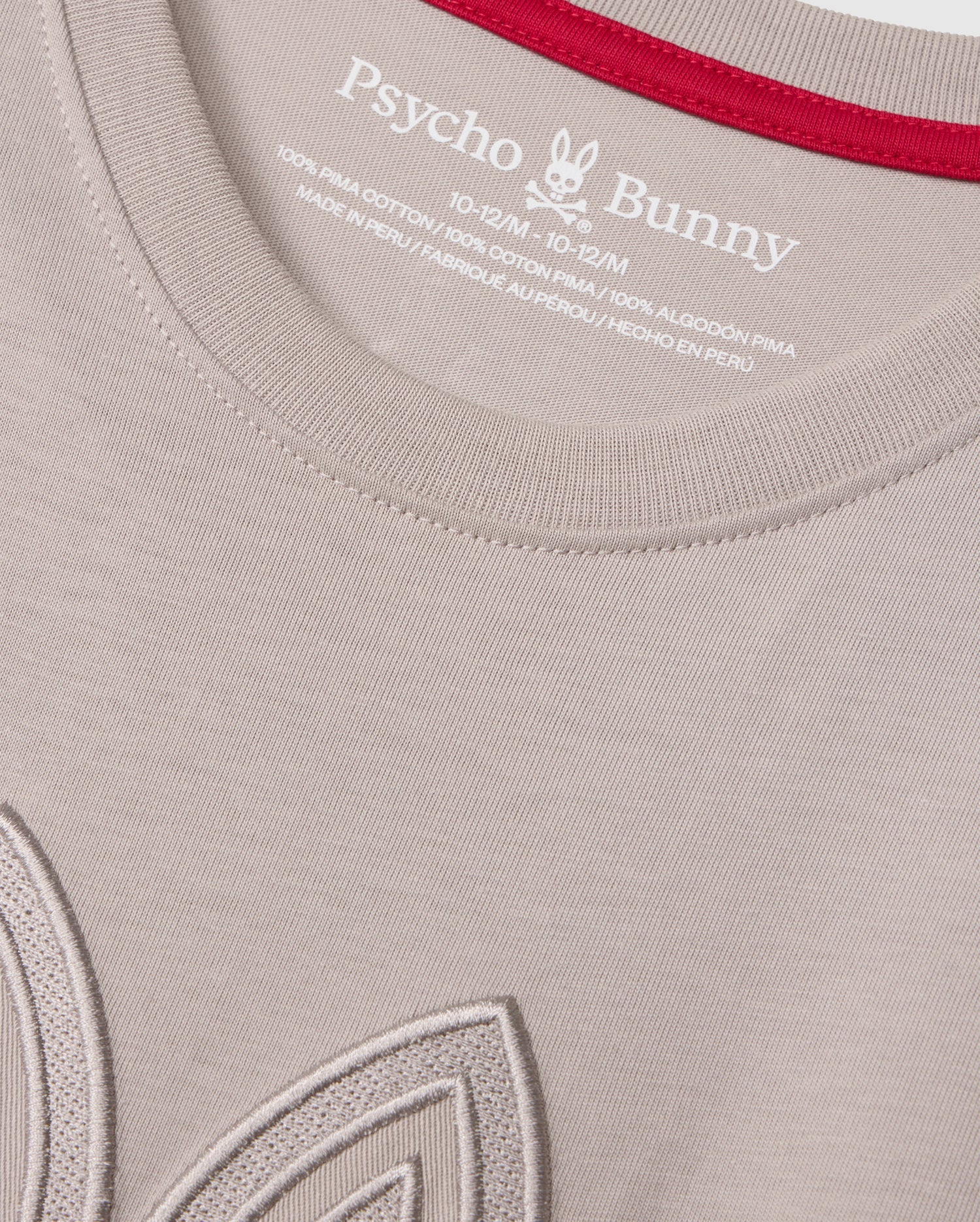 Close-up of a light-colored KIDS MALTON GRAPHIC TEE - B0U795D200 by Psycho Bunny, crafted from soft Pima cotton, featuring the brand's label and size details printed inside the collar. The collar is accented with a red lining, and part of an embroidered design is visible near the bottom left of the image.