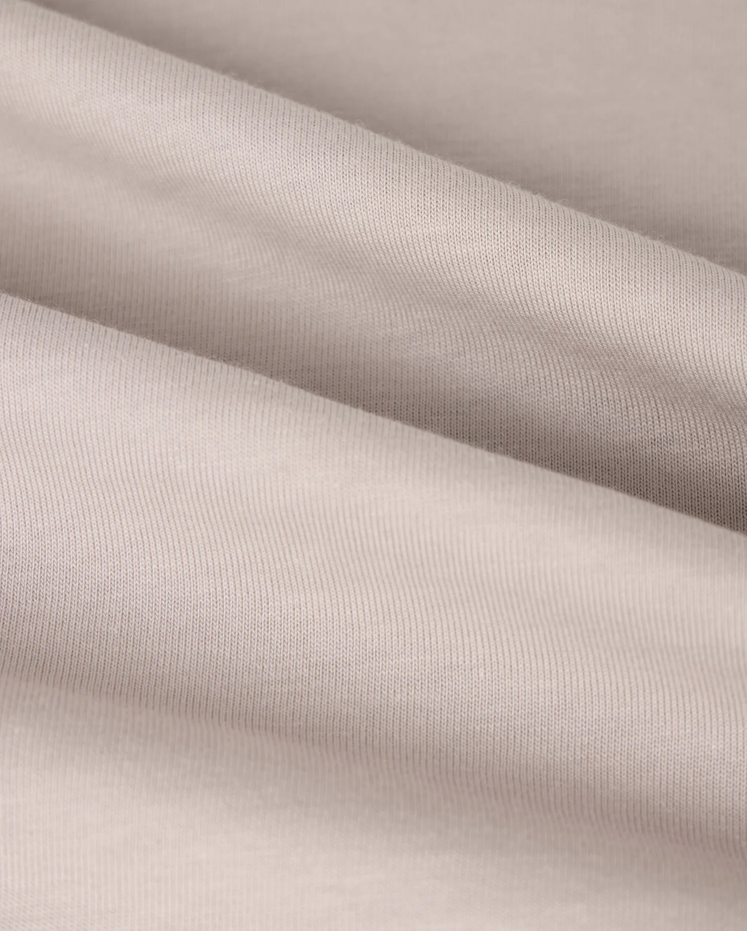 A close-up view of the smooth, beige fabric of the KIDS MALTON GRAPHIC TEE - B0U795D200 from Psycho Bunny shows gentle folds that highlight its soft texture and fine weave. Made from luxurious Pima cotton, the surface appears slightly reflective, creating a subtle play of light and shadow across the material.