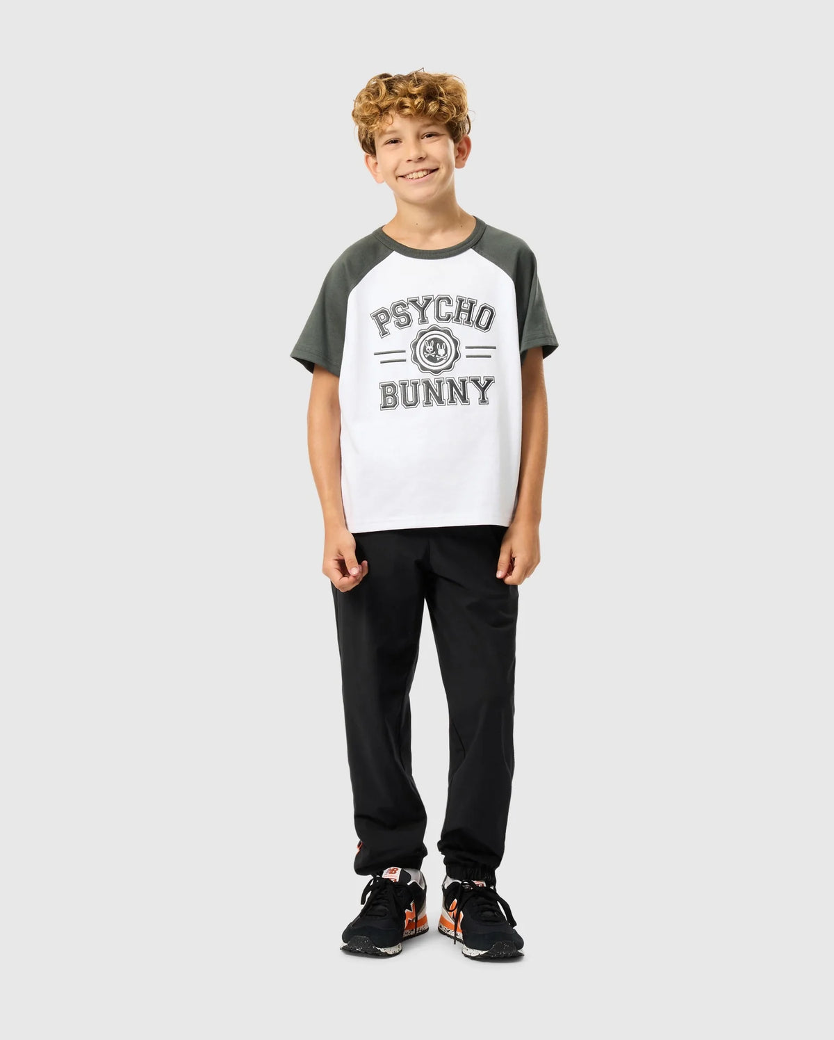 A young boy with curly hair beams in a Psycho Bunny KIDS LONDON RAGLAN GRAPHIC TEE - B0U896D200, known for its premium Pima cotton. He pairs it with black pants and orange-accented sneakers, standing confidently against a plain light gray background.