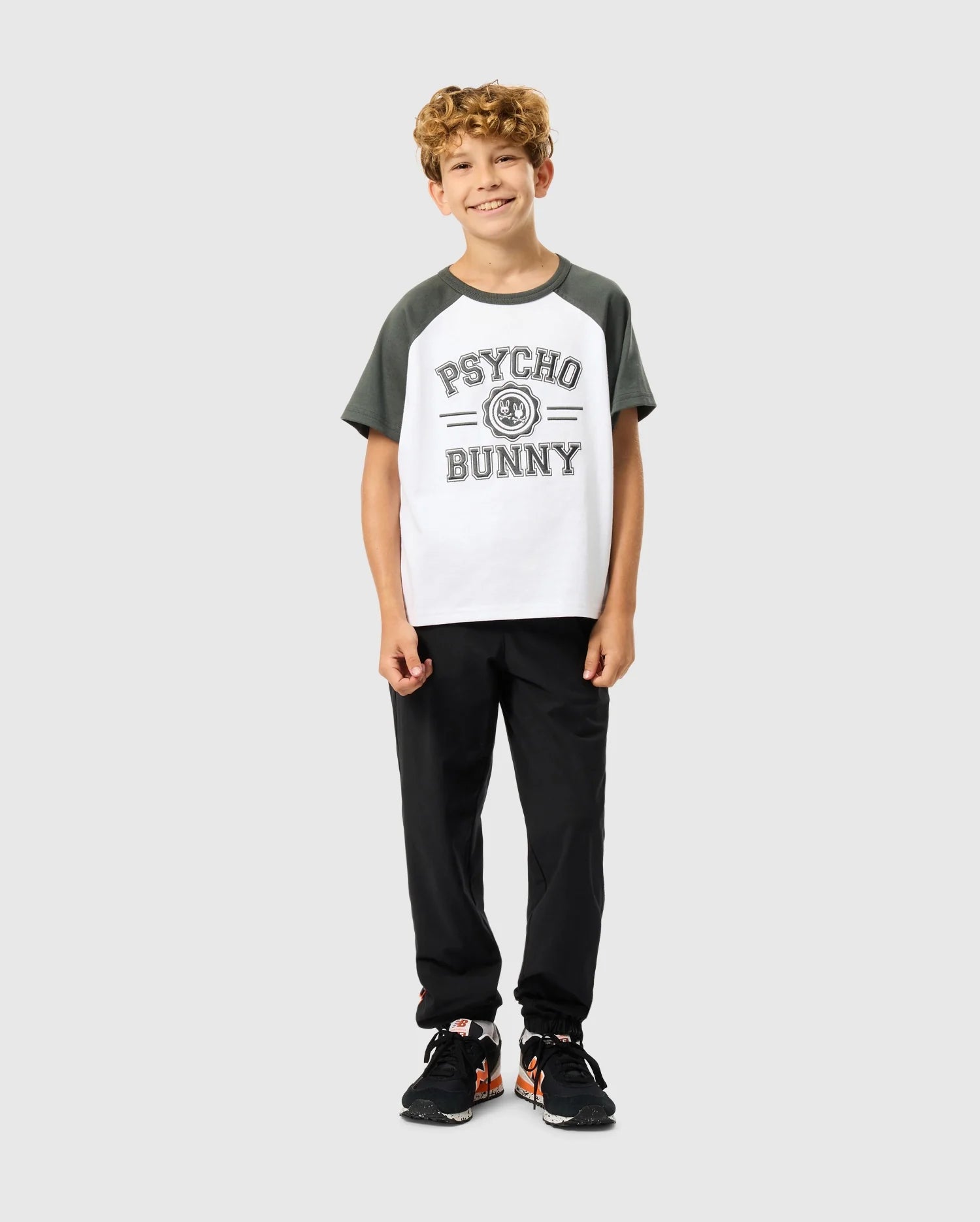 A young boy with curly hair beams in a Psycho Bunny KIDS LONDON RAGLAN GRAPHIC TEE - B0U896D200, known for its premium Pima cotton. He pairs it with black pants and orange-accented sneakers, standing confidently against a plain light gray background.