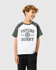 A curly-haired boy smiles confidently in a white and dark green KIDS LONDON RAGLAN GRAPHIC TEE by Psycho Bunny, made from soft Pima cotton, paired with black pants. With one hand in his pocket, he stands against a plain gray background.