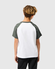 A curly-haired child, wearing a Psycho Bunny KIDS LONDON RAGLAN GRAPHIC TEE in white and dark gray made from soft Pima cotton, paired with dark pants, stands facing away against a plain gray background.