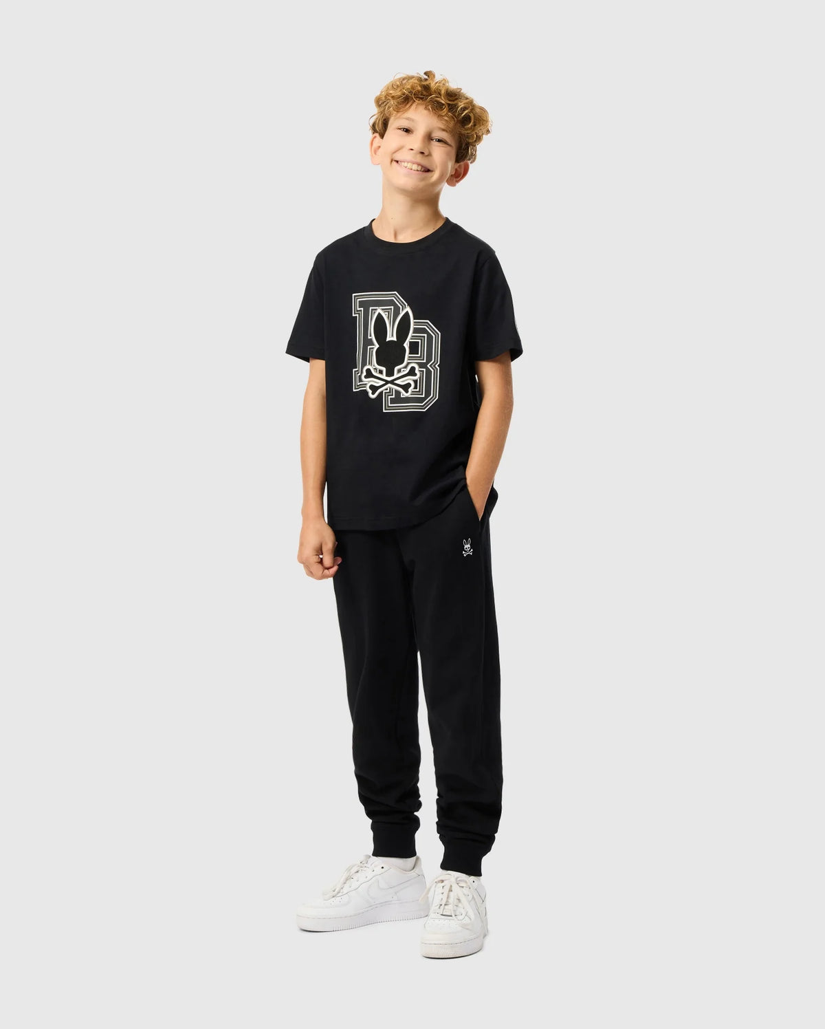 A smiling child with curly hair stands against a plain background, wearing the KIDS ZACK GRAPHIC TEE - B0U897D200 from Psycho Bunny and black sweatpants. With one hand in their pocket and sporting white sneakers, they exude casual charm.
