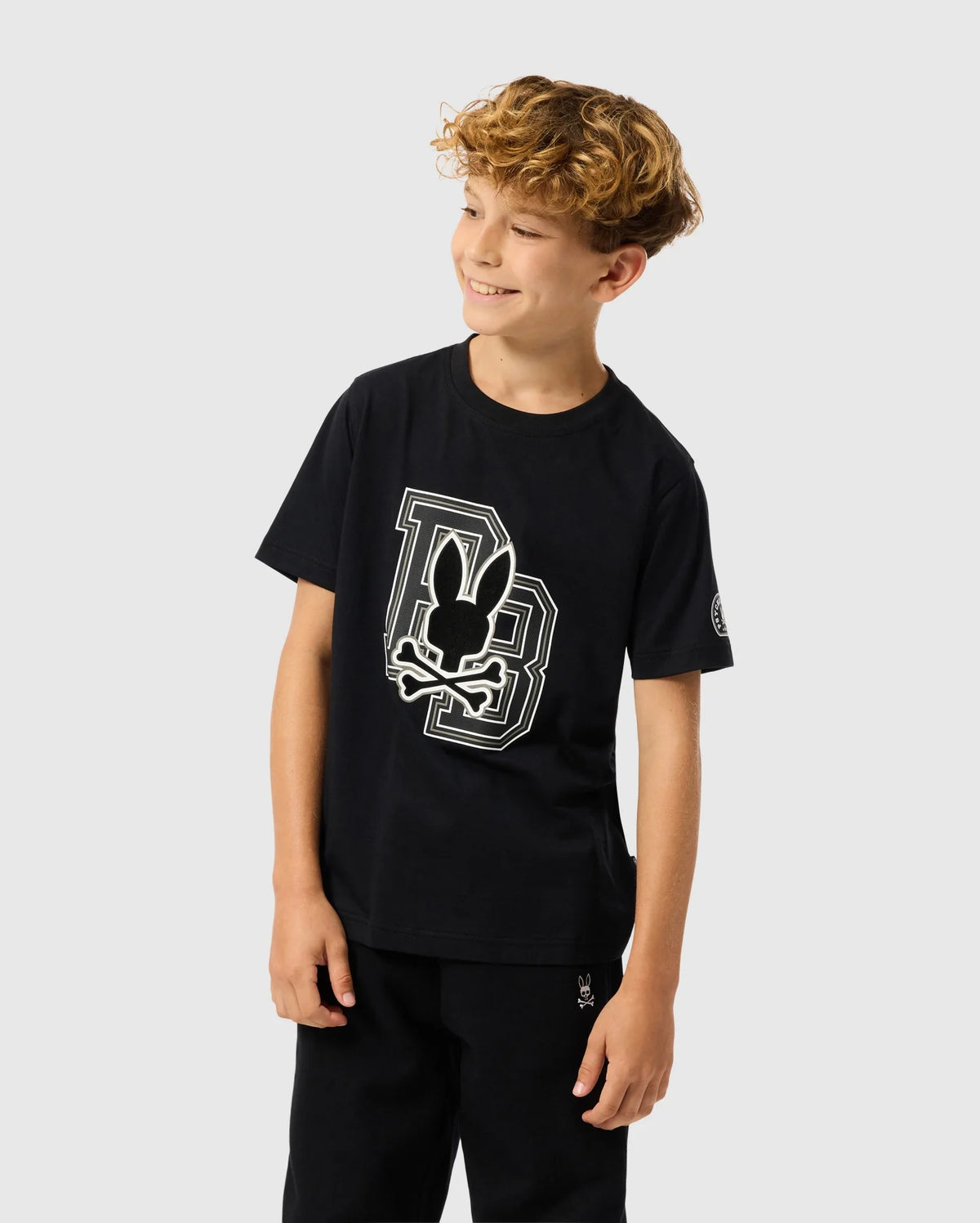 A young boy with curly hair is smiling and looking to the side, wearing a Psycho Bunny Kids Zack Graphic Tee. The black T-shirt features a stylized bunny design with the letters "DB" on soft Pima cotton. The background is plain and light gray.