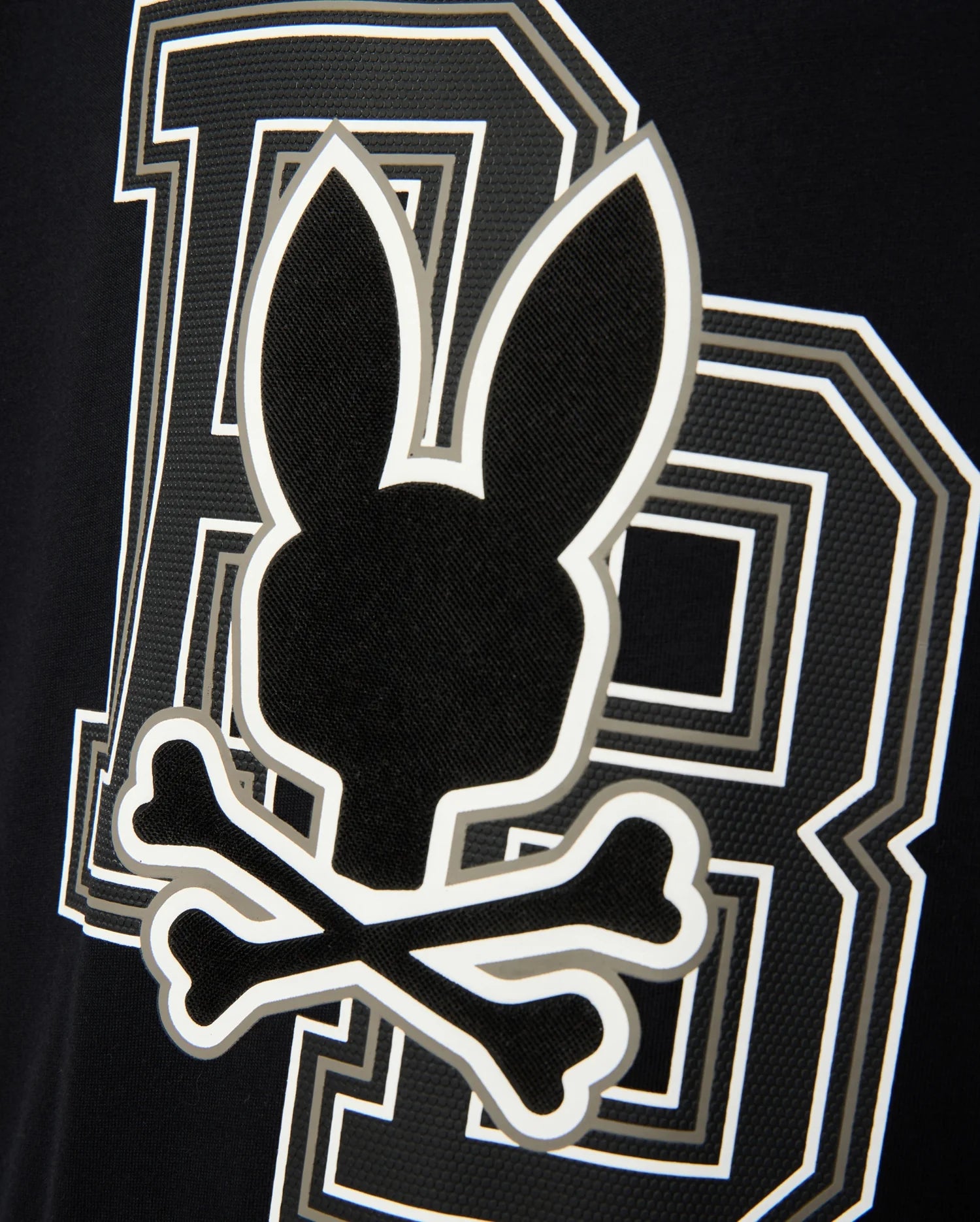 The KIDS ZACK GRAPHIC TEE - B0U897D200 from Psycho Bunny features a black and white design with a Psycho Bunny silhouette above two crossed bones, evoking a skull and crossbones theme, accompanied by a stylized letter "B" in the background. Made from soft Pima cotton, this tee is a striking statement piece.