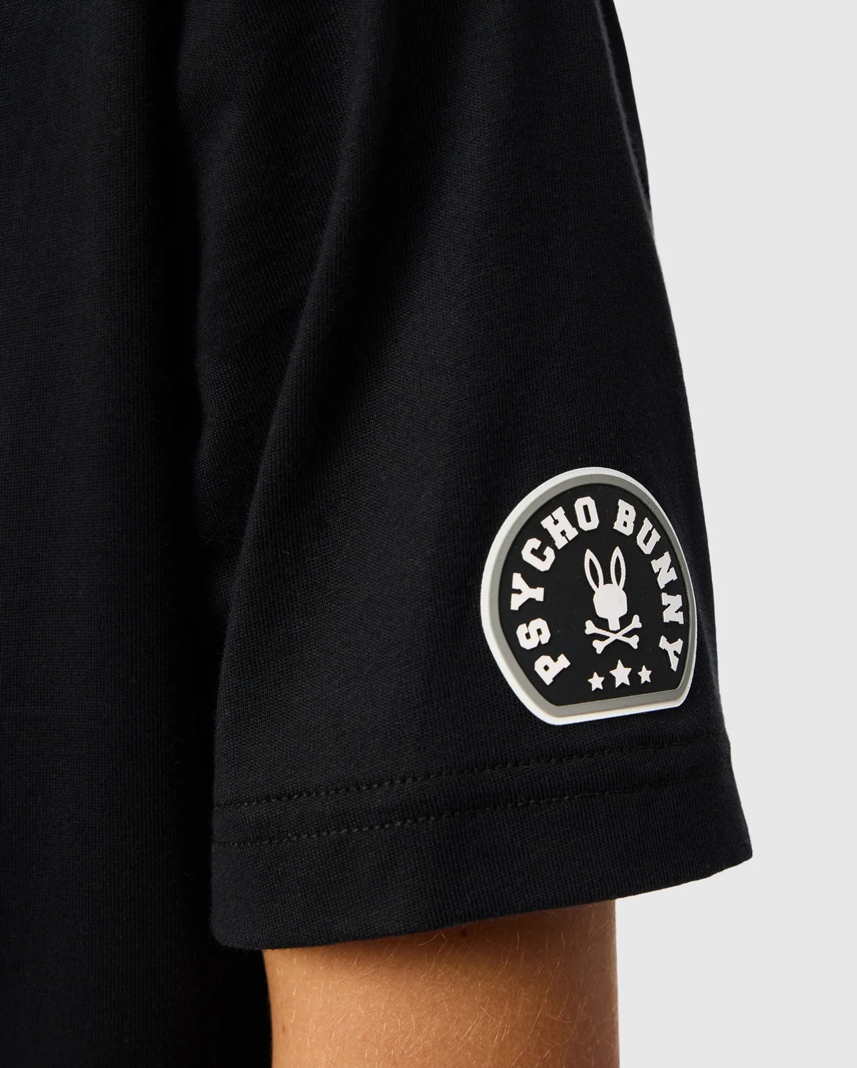 Close-up of a sleeve from the Kids Zack Graphic Tee (B0U897D200) by Psycho Bunny, crafted from luxurious Pima cotton. It features the iconic Psycho Bunny logo—a stylized bunny skull and crossbones topped with stars, encircled by "Psycho Bunny.