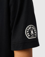 Close-up of a sleeve from the Kids Zack Graphic Tee (B0U897D200) by Psycho Bunny, crafted from luxurious Pima cotton. It features the iconic Psycho Bunny logo—a stylized bunny skull and crossbones topped with stars, encircled by "Psycho Bunny.