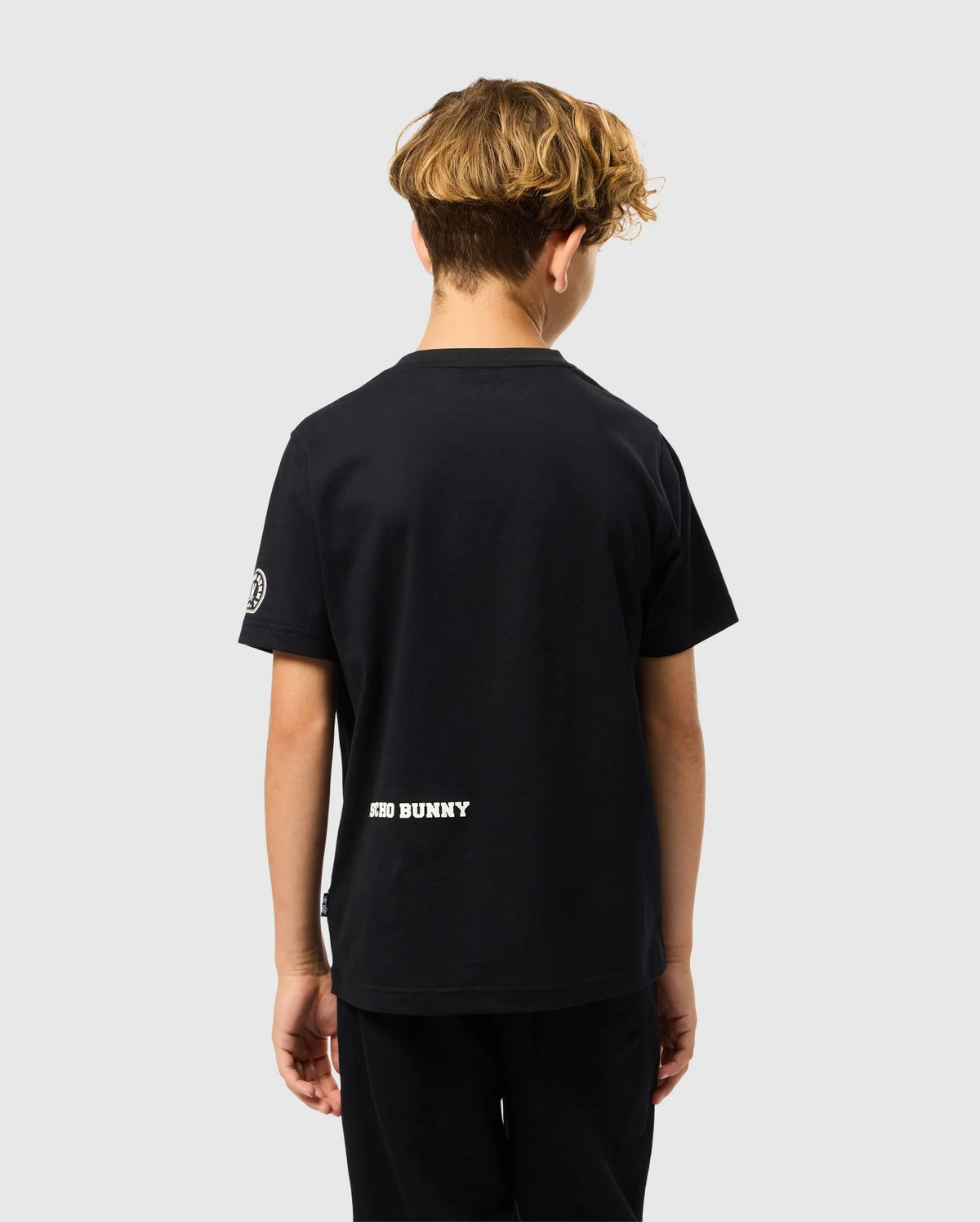 A young individual with curly hair faces away from the camera, clad in a black KIDS ZACK GRAPHIC TEE - B0U897D200 and pants. Made from soft Pima cotton, this Psycho Bunny shirt prominently displays "BAD BUNNY" near the hem against a simple, light gray backdrop.