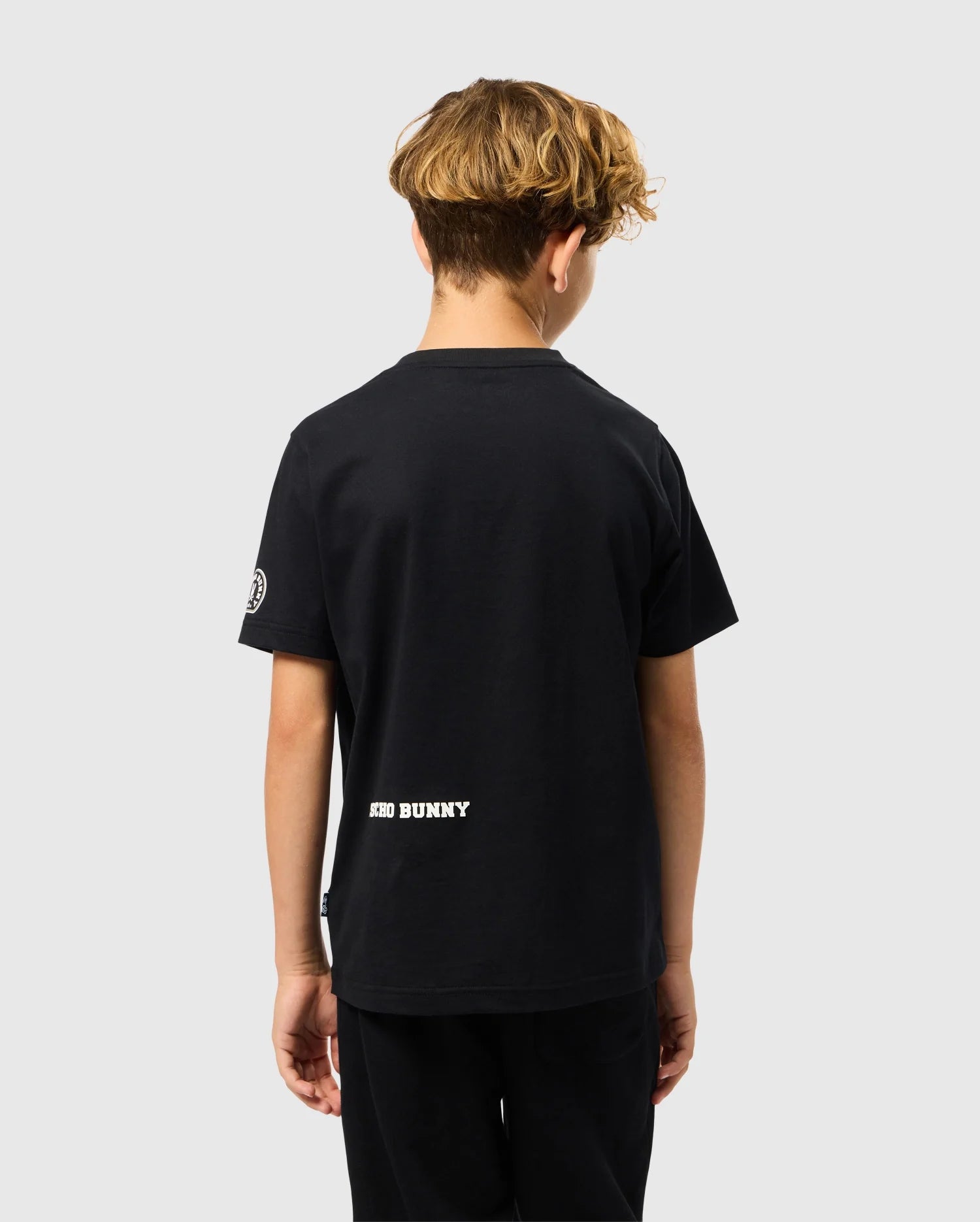 A young individual with curly hair faces away from the camera, clad in a black KIDS ZACK GRAPHIC TEE - B0U897D200 and pants. Made from soft Pima cotton, this Psycho Bunny shirt prominently displays "BAD BUNNY" near the hem against a simple, light gray backdrop.