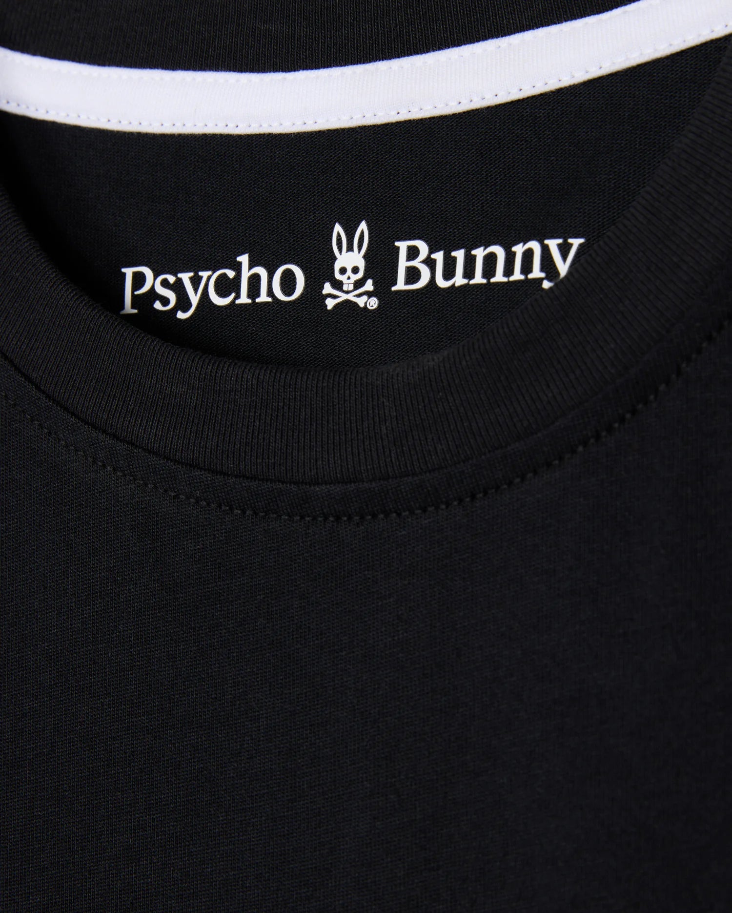 Close-up of the KIDS ZACK GRAPHIC TEE (B0U897D200) from Psycho Bunny, featuring a black Pima cotton shirt collar with the brand's iconic bunny head and crossbones logo, accented by white stitching along the neckline.