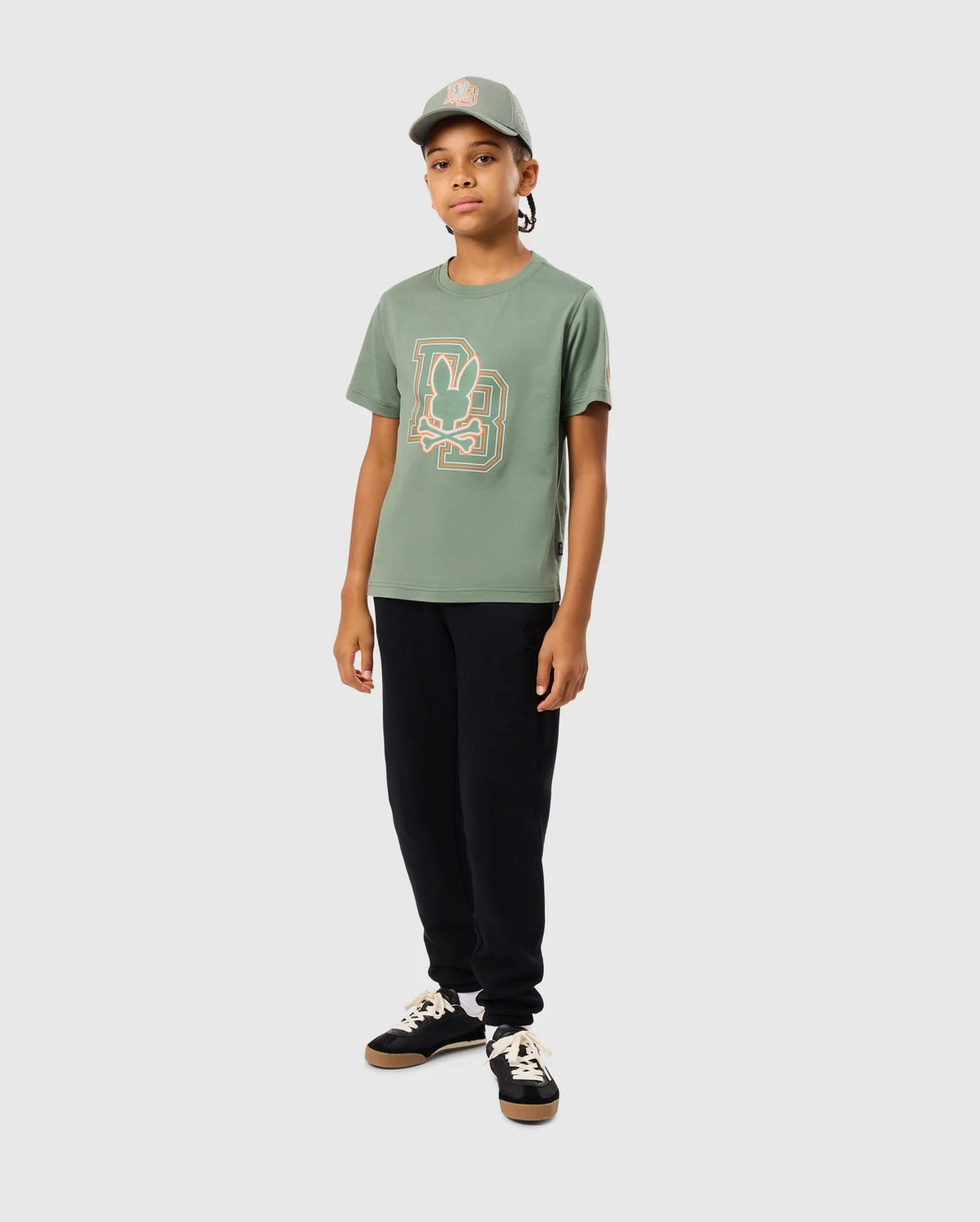 A young person stands against a plain background, wearing the KIDS ZACK GRAPHIC TEE - B0U897D200 in green by Psycho Bunny. They pair it with black pants, a matching green cap, and black sneakers with white laces. Their long hair flows as they gaze confidently toward the camera.