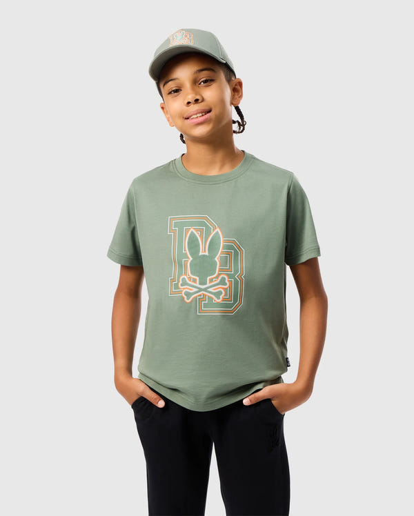A young person wearing the Kids Zack Graphic Tee - B0U897D200 by Psycho Bunny in green, made from Pima cotton, with a matching green cap. They stand with their hands in the pockets of black pants against a light gray background.