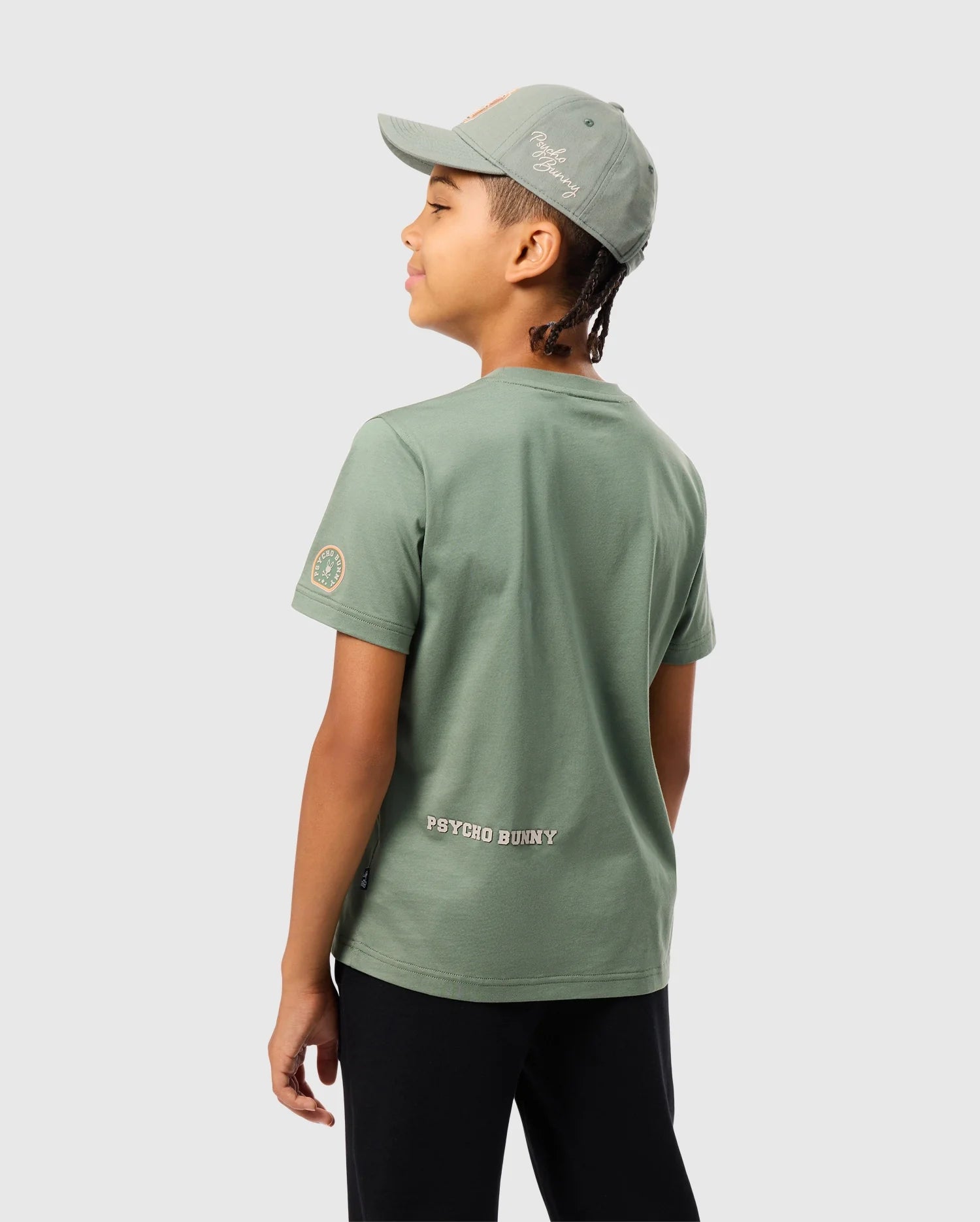 A young child is dressed in a green KIDS ZACK GRAPHIC TEE (B0U897D200) from Psycho Bunny, made from soft Pima cotton, paired with a matching cap. They face slightly away to display the back design of the tee. The child has short braided hair and wears black pants against a plain gray background.