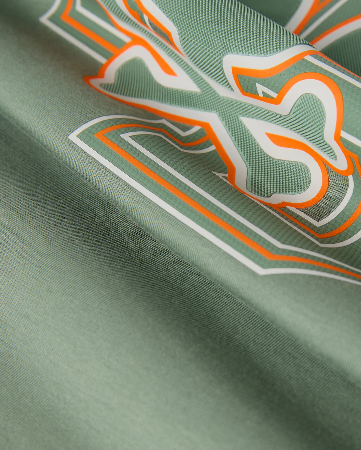 A detailed view of the KIDS ZACK GRAPHIC TEE - B0U897D200 by Psycho Bunny shows green Pima cotton fabric adorned with an abstract design featuring white, orange, and dark green outlines in a bold, dynamic pattern. The texture is smooth and slightly shiny.