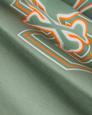 A detailed view of the KIDS ZACK GRAPHIC TEE - B0U897D200 by Psycho Bunny shows green Pima cotton fabric adorned with an abstract design featuring white, orange, and dark green outlines in a bold, dynamic pattern. The texture is smooth and slightly shiny.