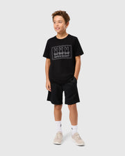 A young boy with curly hair stands against a plain background, wearing the KIDS TACOMA GRAPHIC TEE - B0U909D200 by Psycho Bunny, made of soft Pima cotton and featuring three cartoon rabbits labeled "PHOTOGRAPHY." He is also wearing black shorts, white socks, and beige sneakers.
