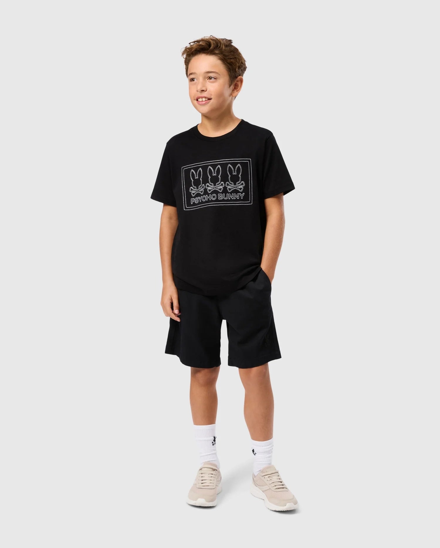 A young boy with curly hair stands against a plain background, wearing the KIDS TACOMA GRAPHIC TEE - B0U909D200 by Psycho Bunny, made of soft Pima cotton and featuring three cartoon rabbits labeled "PHOTOGRAPHY." He is also wearing black shorts, white socks, and beige sneakers.