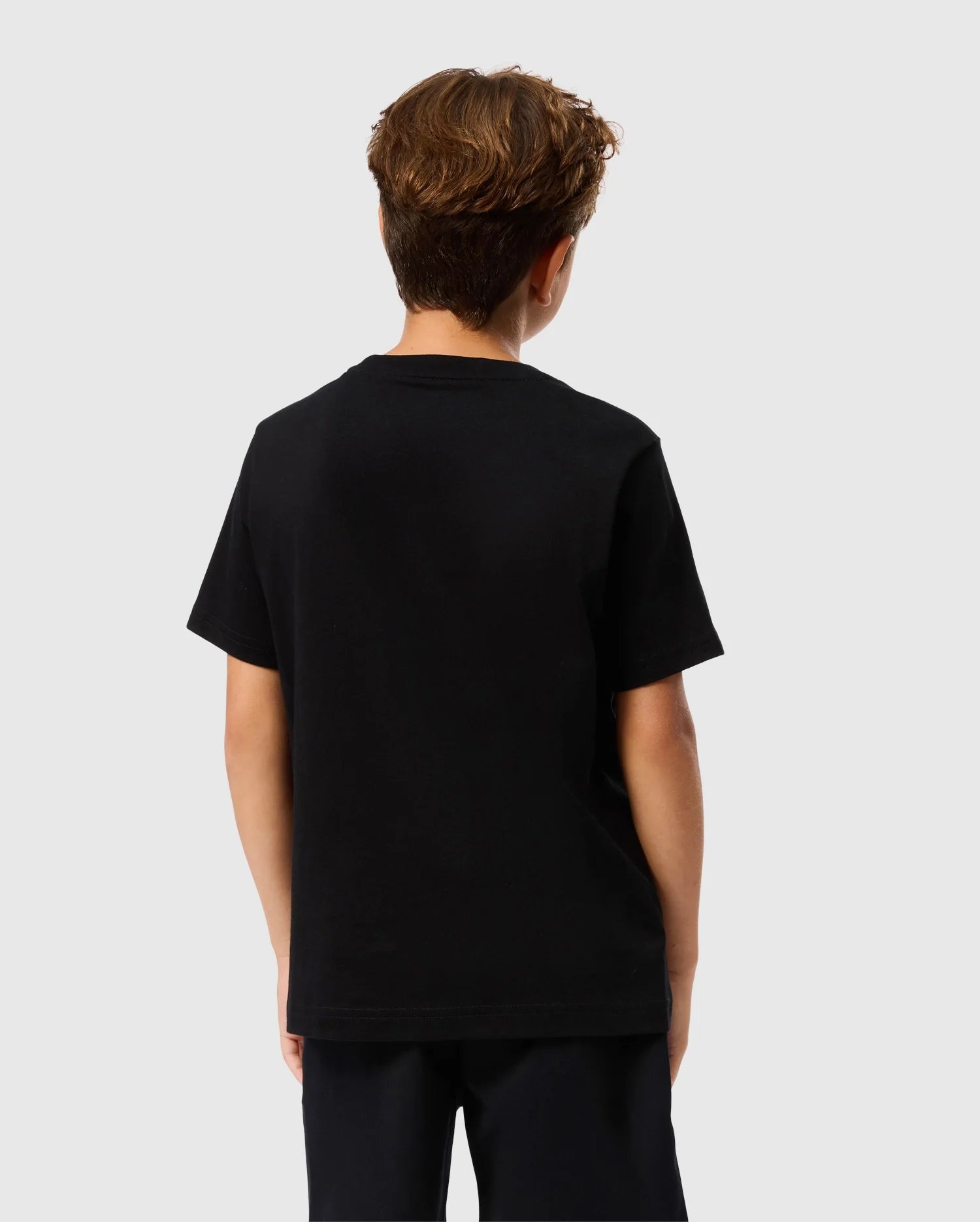 A young person with short brown hair is wearing the KIDS TACOMA GRAPHIC TEE - B0U909D200 by Psycho Bunny and black pants featuring subtle embroidered details, as they face away from the camera against a light gray background.