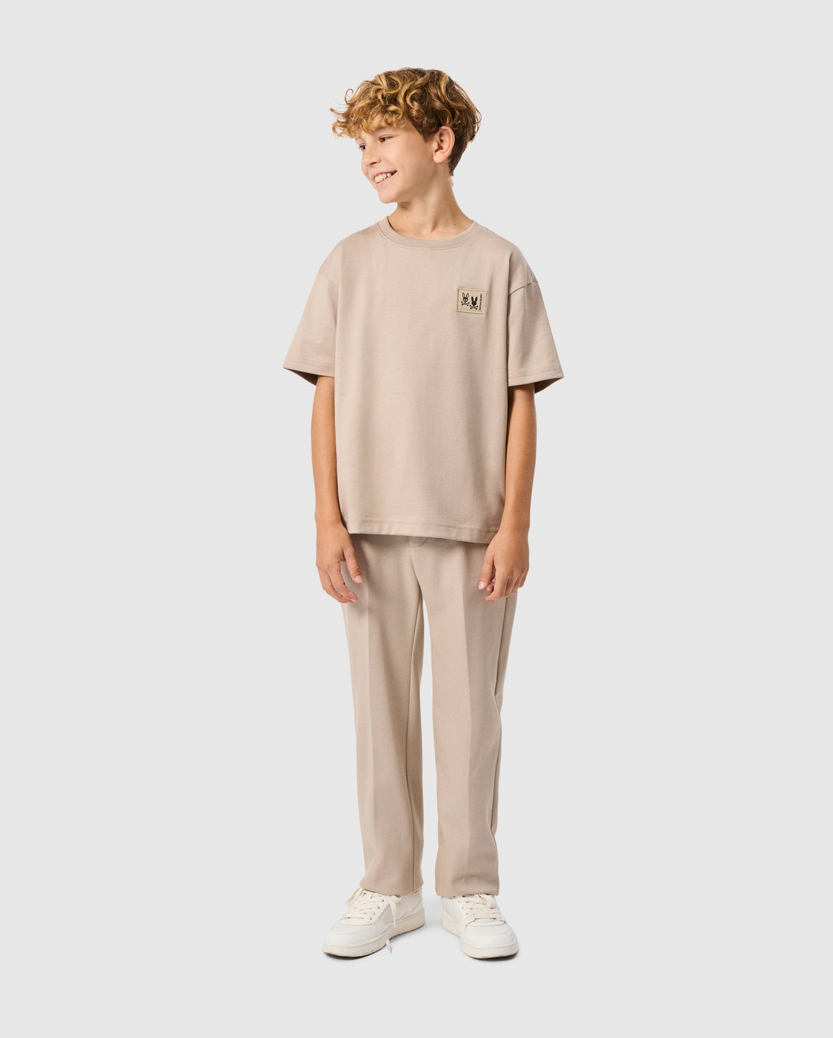 A young boy with curly hair smiles confidently, dressed in a beige Psycho Bunny KIDS MONROE HEAVY WEIGHT RELAXED FIT TEE and pants, paired with white sneakers, against a plain white background.