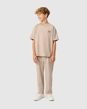 A young boy with curly hair smiles confidently, dressed in a beige Psycho Bunny KIDS MONROE HEAVY WEIGHT RELAXED FIT TEE and pants, paired with white sneakers, against a plain white background.
