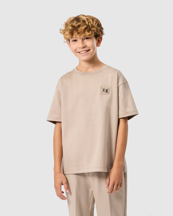 A curly blond boy smiles wearing a Psycho Bunny KIDS MONROE HEAVY WEIGHT RELAXED FIT TEE in beige with matching pants, featuring a small chest logo, against a plain white background.