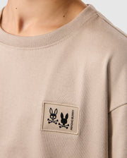 The Psycho Bunny KIDS MONROE HEAVY WEIGHT RELAXED FIT TEE features a beige Pima cotton design with a rectangular patch displaying two black cartoon rabbits and crossed bones, alongside the "ROCKO & BUNNY" text, exuding a laid-back vibe similar to a men's relaxed fit tee.