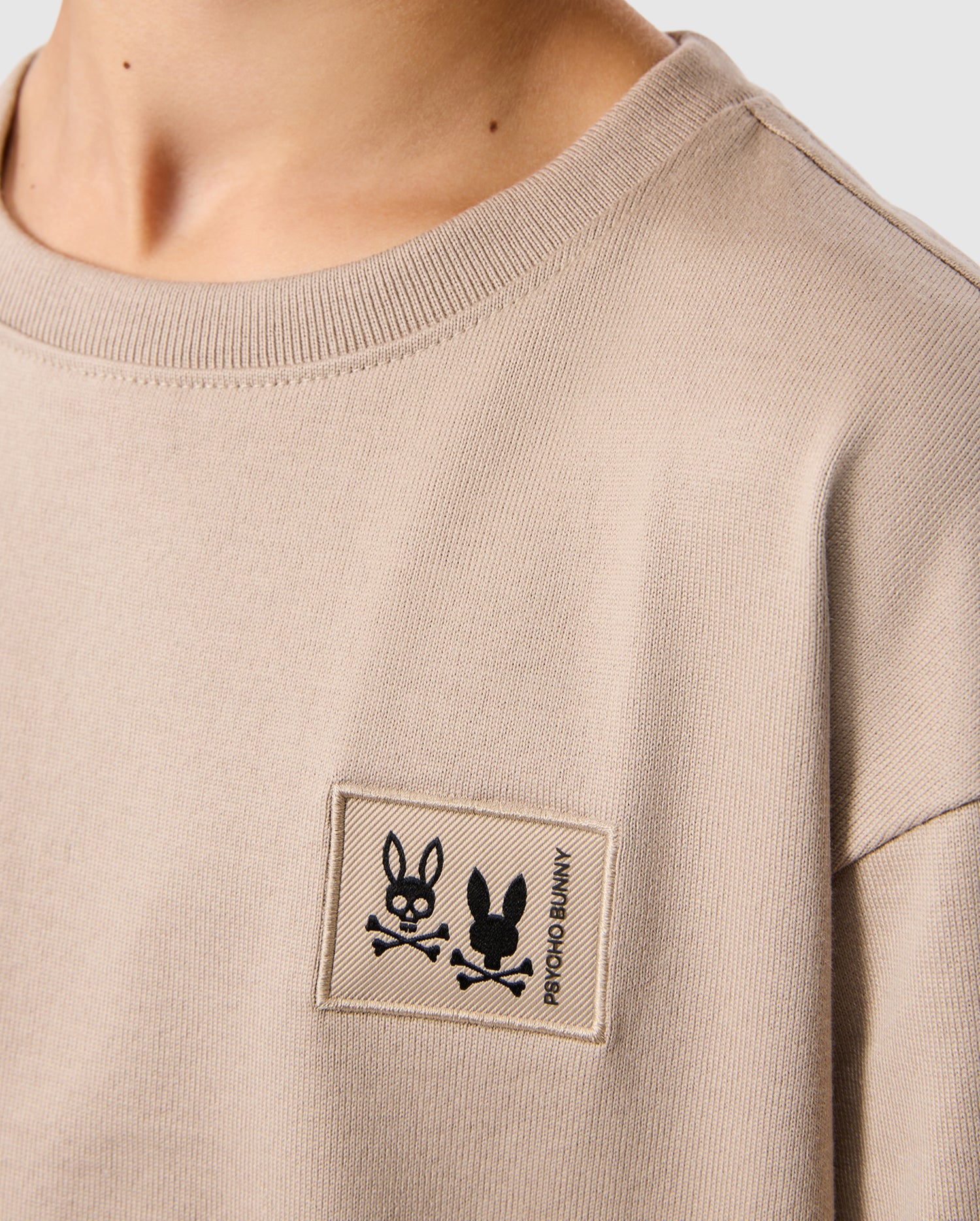 The Psycho Bunny KIDS MONROE HEAVY WEIGHT RELAXED FIT TEE features a beige Pima cotton design with a rectangular patch displaying two black cartoon rabbits and crossed bones, alongside the "ROCKO & BUNNY" text, exuding a laid-back vibe similar to a men's relaxed fit tee.