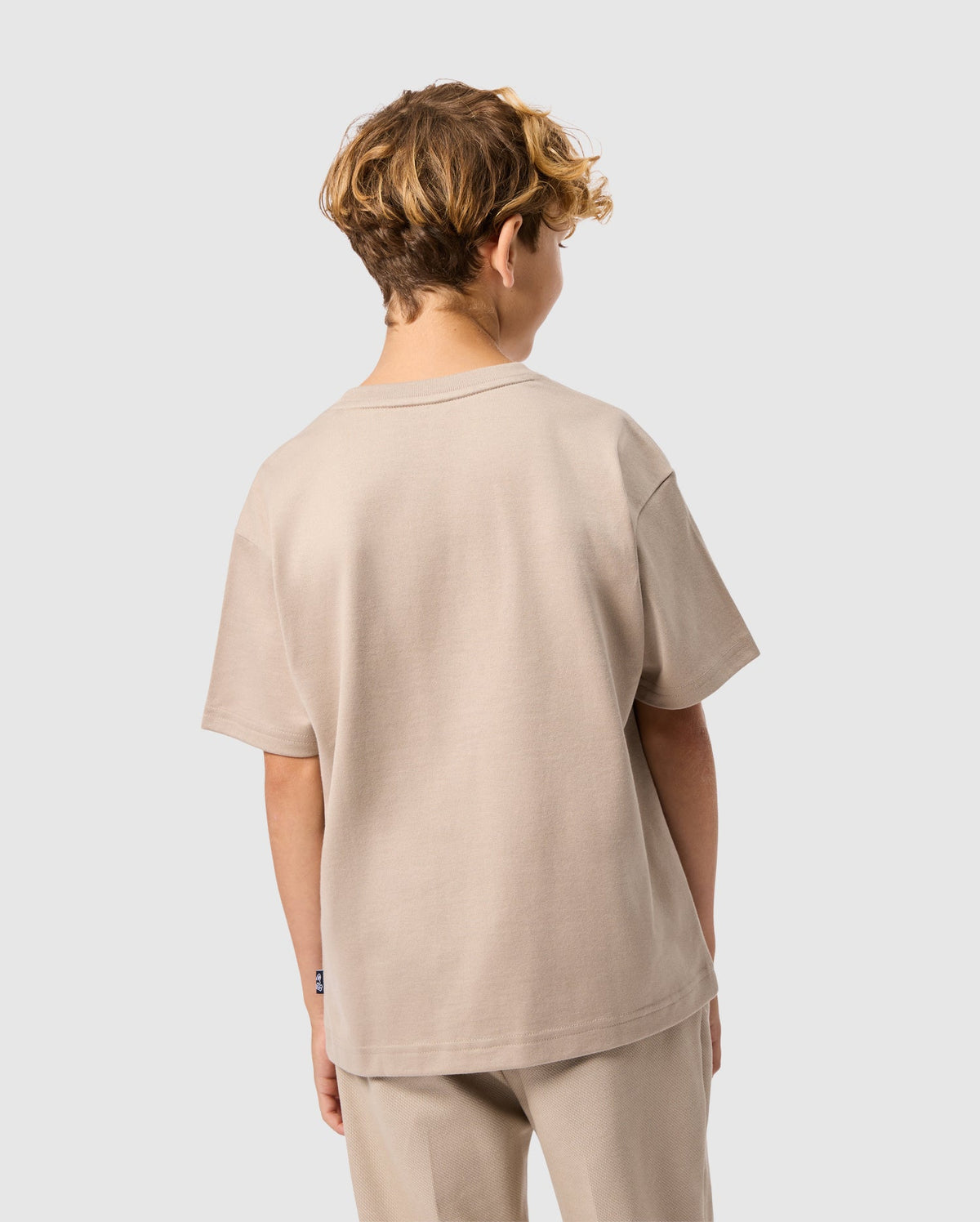 A child with curly hair wears a Psycho Bunny KIDS MONROE HEAVY WEIGHT RELAXED FIT TEE in light beige and matching pants, standing back to the camera against a plain, light gray background, showcasing their casual style.
