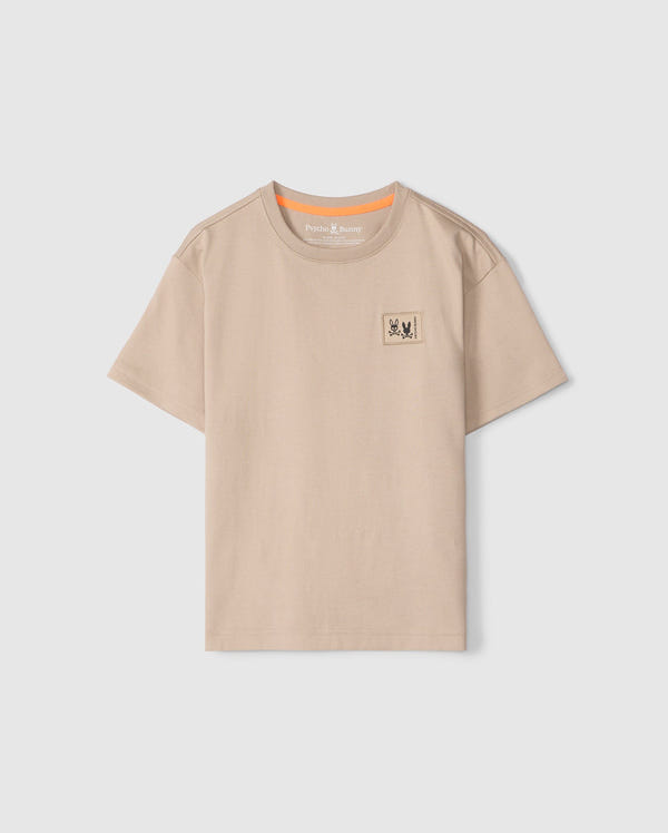 The KIDS MONROE HEAVY WEIGHT RELAXED FIT TEE by Psycho Bunny is made from soft Pima cotton, featuring a beige hue, short sleeves, and a round neckline. It includes a small printed patch with two stylized animals on the upper left chest and an orange accent inside the collar.