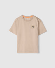 The KIDS MONROE HEAVY WEIGHT RELAXED FIT TEE by Psycho Bunny is made from soft Pima cotton, featuring a beige hue, short sleeves, and a round neckline. It includes a small printed patch with two stylized animals on the upper left chest and an orange accent inside the collar.