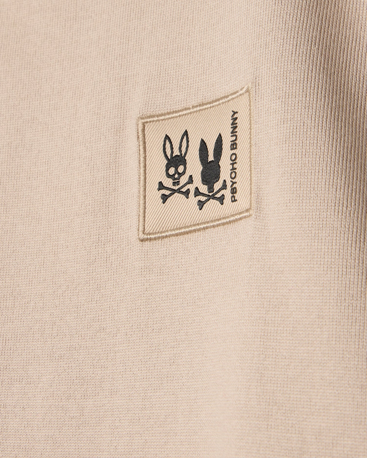 The KIDS MONROE HEAVY WEIGHT RELAXED FIT TEE in beige features a small square patch with black rabbit skull illustrations above crossed bones, and "Psycho Bunny" printed vertically next to the patch. Crafted from premium Pima cotton, it offers a casual style.