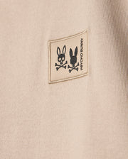 The KIDS MONROE HEAVY WEIGHT RELAXED FIT TEE in beige features a small square patch with black rabbit skull illustrations above crossed bones, and "Psycho Bunny" printed vertically next to the patch. Crafted from premium Pima cotton, it offers a casual style.