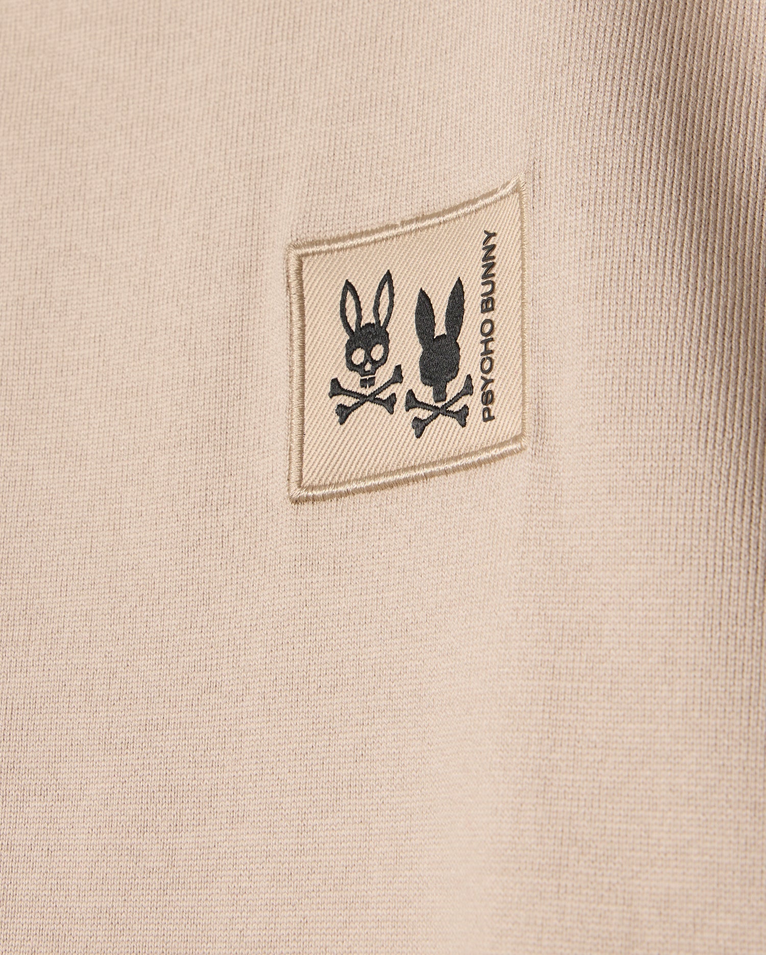 The KIDS MONROE HEAVY WEIGHT RELAXED FIT TEE in beige features a small square patch with black rabbit skull illustrations above crossed bones, and "Psycho Bunny" printed vertically next to the patch. Crafted from premium Pima cotton, it offers a casual style.