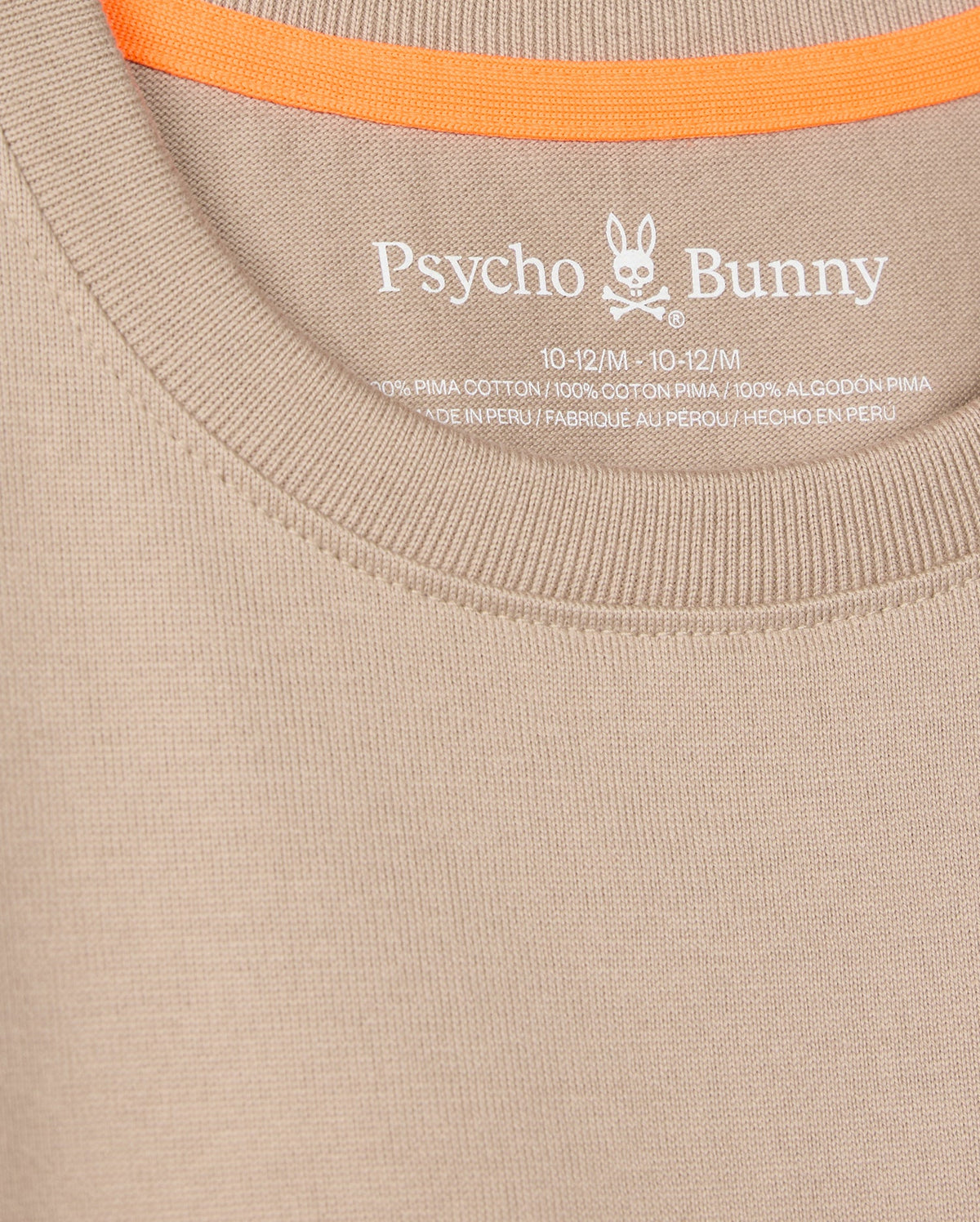 The close-up shows a beige Psycho Bunny tee (KIDS MONROE HEAVY WEIGHT RELAXED FIT TEE - B0U910D200) with the logo and brand elegantly printed inside the neckline. Made of Pima cotton, it has an orange trim on the inner back collar for casual style.