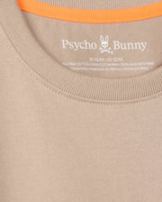 The close-up shows a beige Psycho Bunny tee (KIDS MONROE HEAVY WEIGHT RELAXED FIT TEE - B0U910D200) with the logo and brand elegantly printed inside the neckline. Made of Pima cotton, it has an orange trim on the inner back collar for casual style.