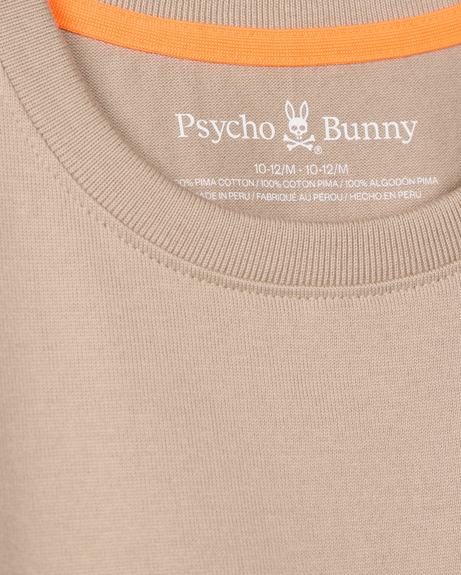 The close-up shows a beige Psycho Bunny tee (KIDS MONROE HEAVY WEIGHT RELAXED FIT TEE - B0U910D200) with the logo and brand elegantly printed inside the neckline. Made of Pima cotton, it has an orange trim on the inner back collar for casual style.