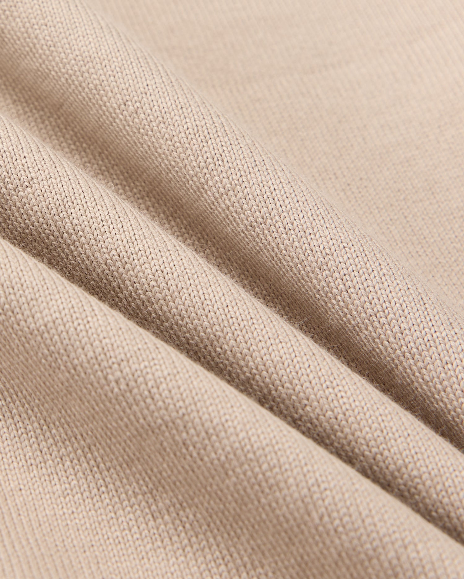 Close-up of beige fabric, showcasing a textured knit pattern with three soft folds that highlight its flexibility and smooth surface. The play of light and shadow defines the fine weave details, perfect for Psycho Bunny's KIDS MONROE HEAVY WEIGHT RELAXED FIT TEE, B0U910D200.