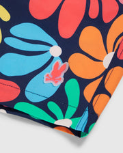 A close-up image showcases the colorful floral-patterned fabric of the KIDS MENTZ SWIM TRUNK - B0W320B2SW by Psycho Bunny, adorned with large, vibrant flowers in red, blue, green, and orange against a dark background. A small pink and white rabbit logo with crossed bones is affixed near the seam on these quick-dry swim trunks.