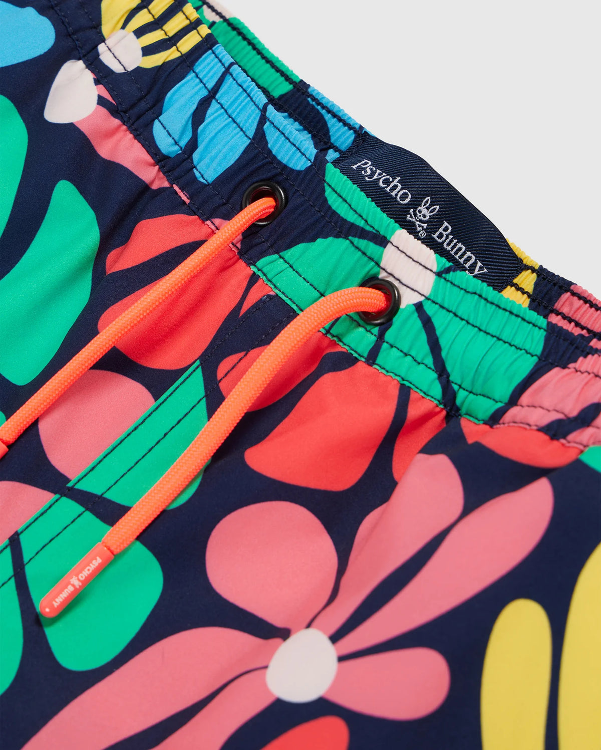 Close-up image of colorful KIDS MENTZ SWIM TRUNK (B0W320B2SW) featuring a mix of vibrant floral patterns in blue, purple, green, yellow, and pink. The waistband includes orange drawstrings and a black tag with "Psycho Bunny" branding. The material appears lightweight and suitable for water activities.