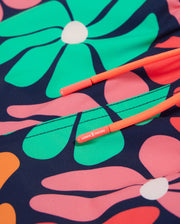 Close-up image of Psycho Bunny's KIDS MENTZ SWIM TRUNK - B0W320B2SW, showcasing a vibrant fabric featuring a floral pattern with large pink, green, and orange flowers on a dark blue background. Bright orange drawstrings with small tags are visible, suggesting this is part of the quick-dry swim trunk design.